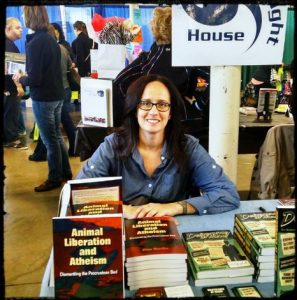 Photo of Kim behind stacks of books on a table. Freethought House banner visible in background.