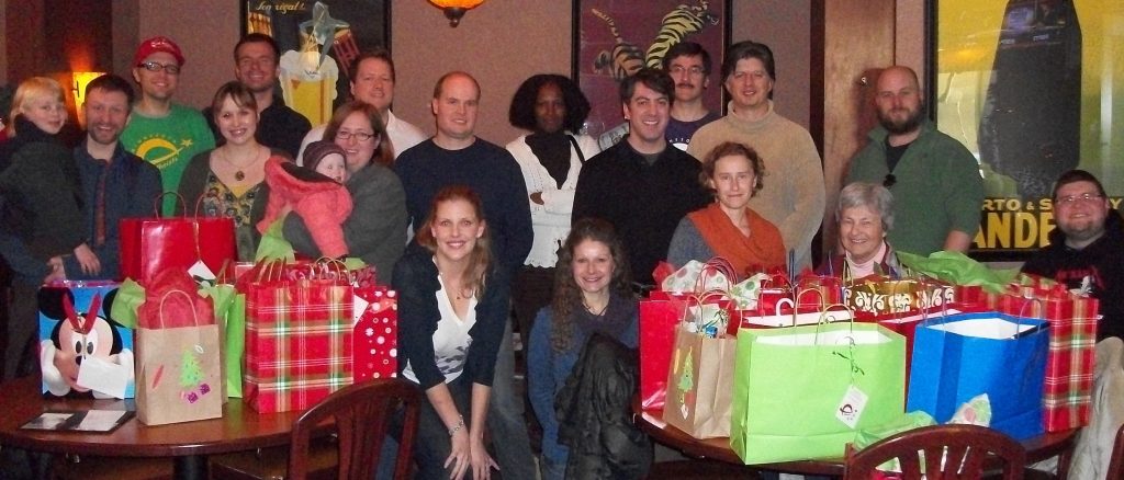 Photo of about 20 people standing behind tables full of wrapped presents.